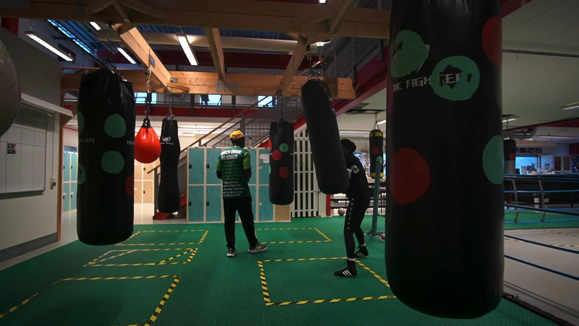 En boxningslokal med hängande säckar på grönt golv, där en boxare tränar och en tränare står bredvid.
