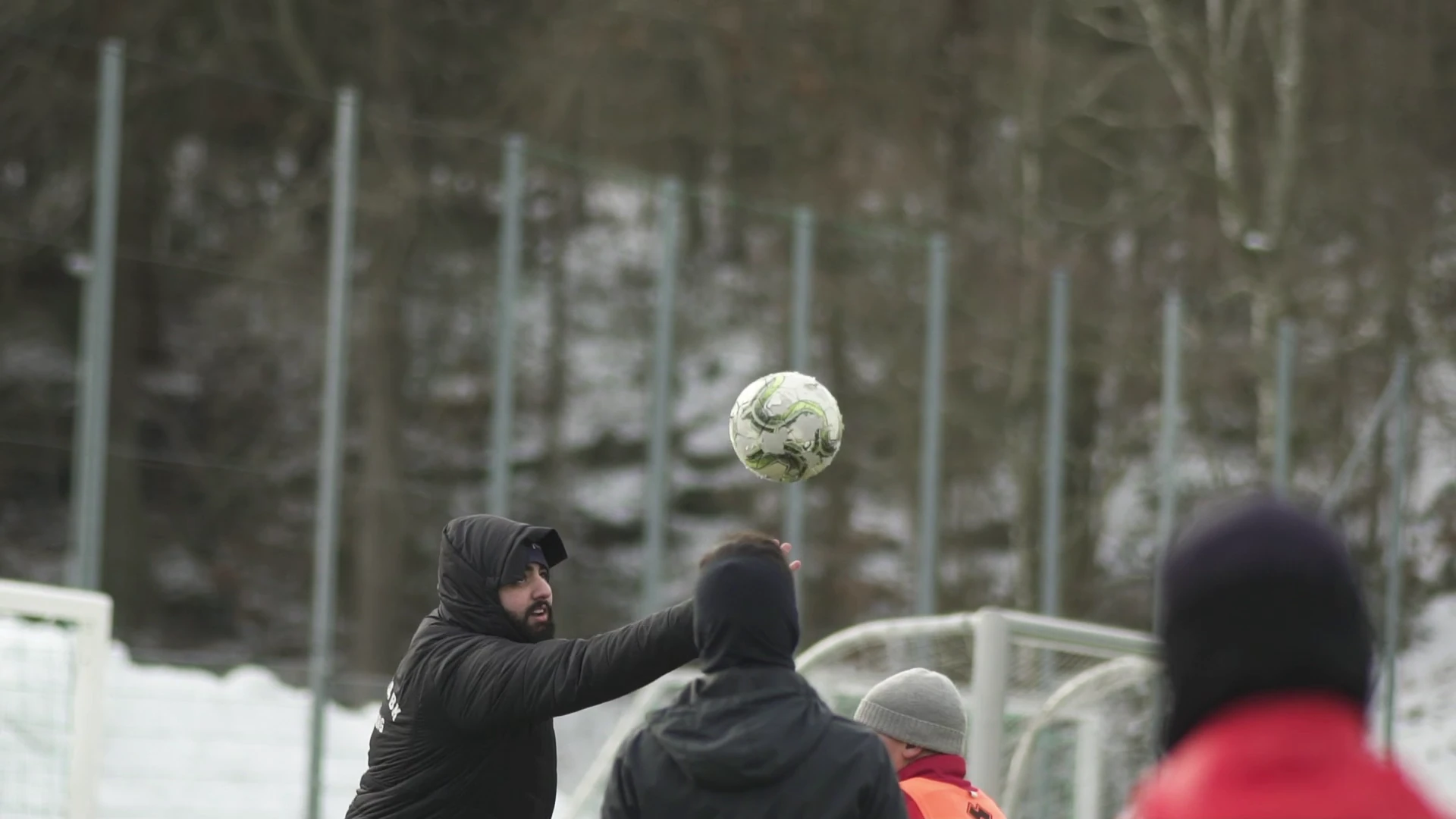 En fotboll i luften under träning på en snöig plan, där en man i svart vinterjacka sträcker ut armen.