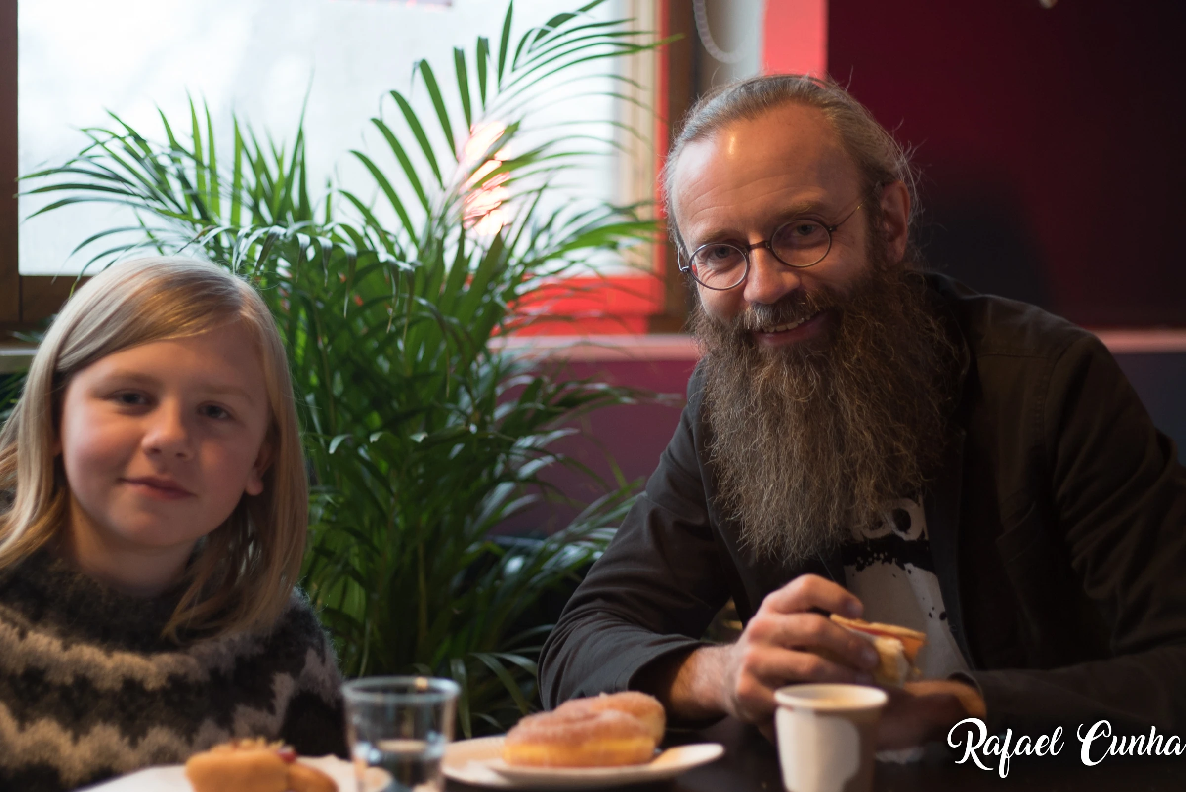 A child and a man with a long beard sit at a café table and smile at the camera.