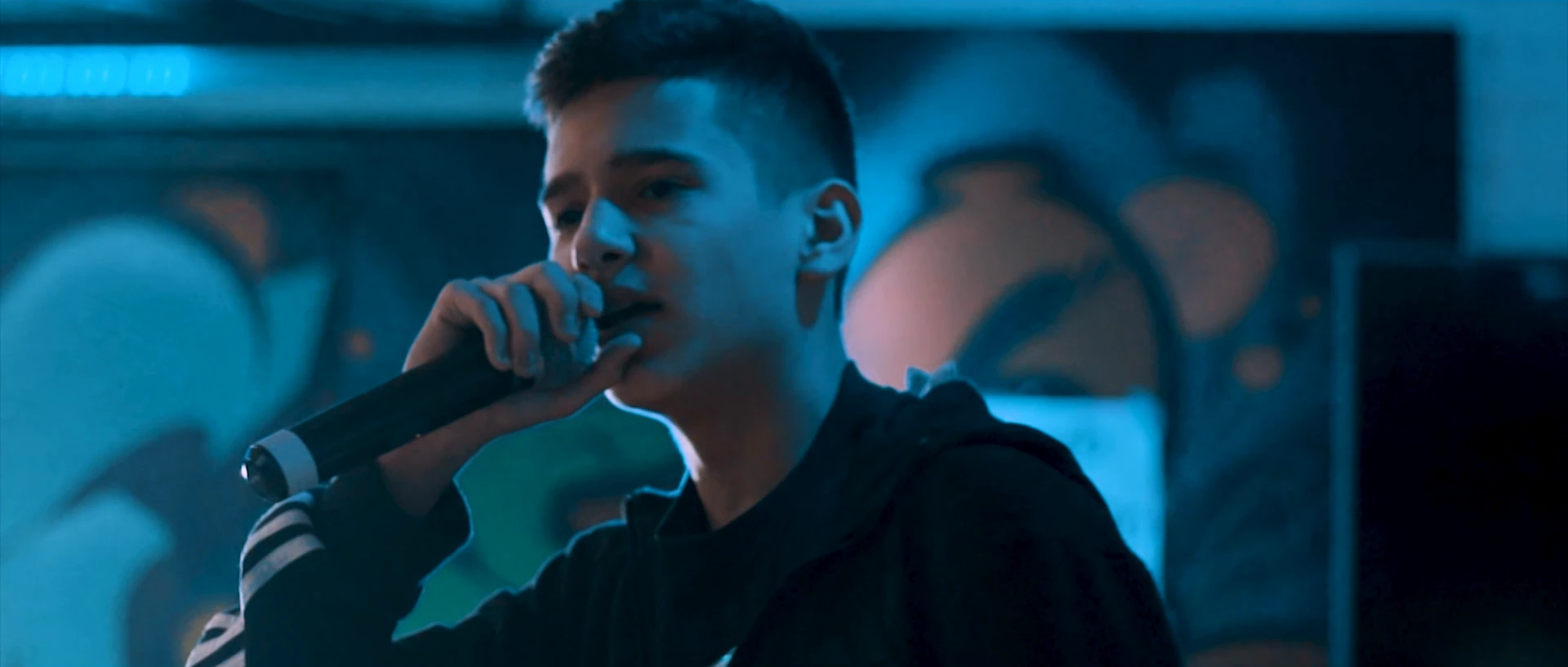 A young man raps into a microphone in front of a graffiti wall in blue light.