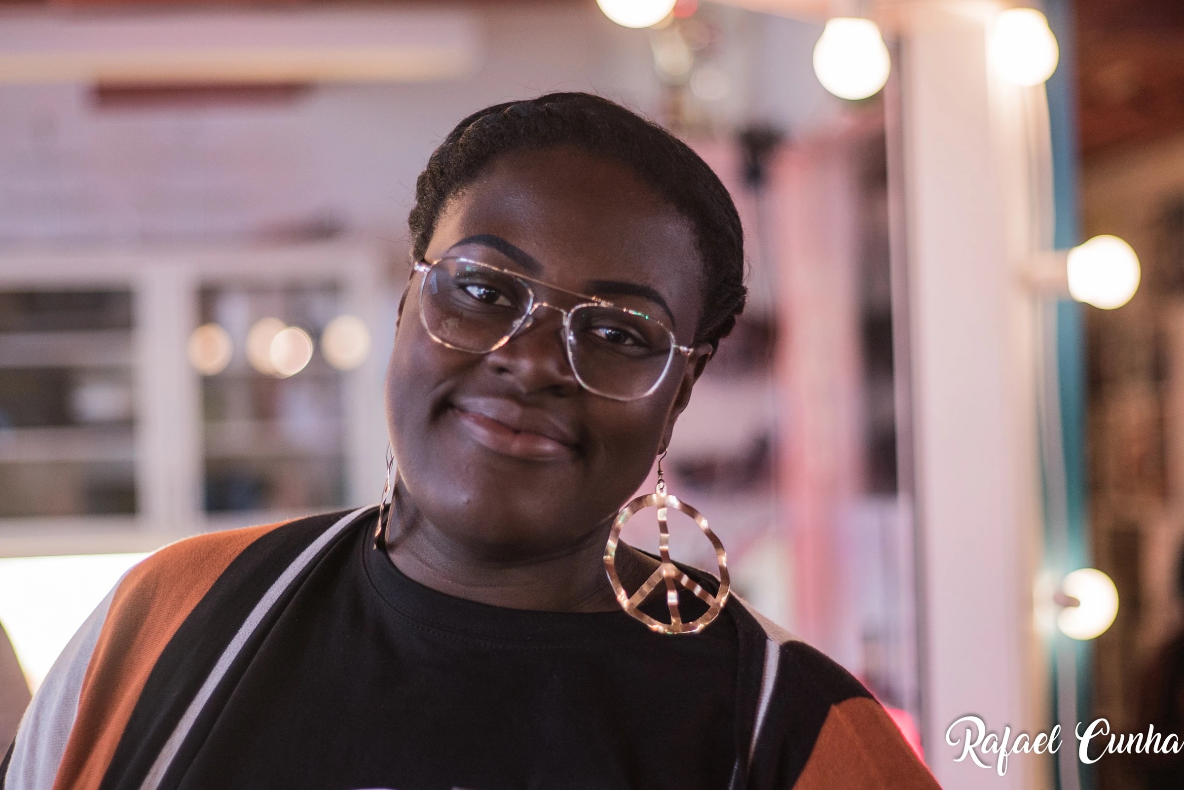 A woman in large glasses and an earring shaped like a peace symbol tilts her head and smiles.