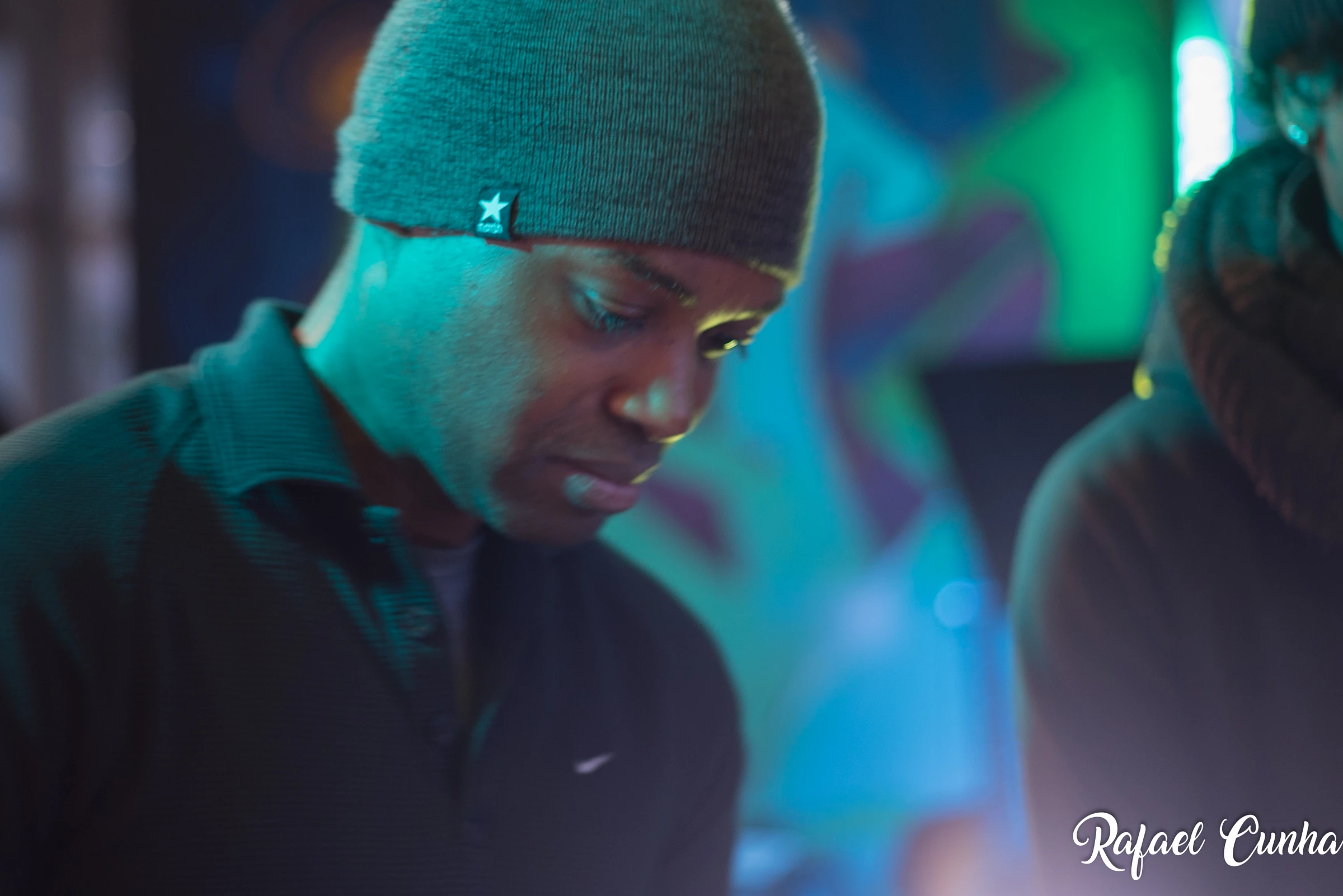 A man in a beanie looks down, lit by green and blue light.