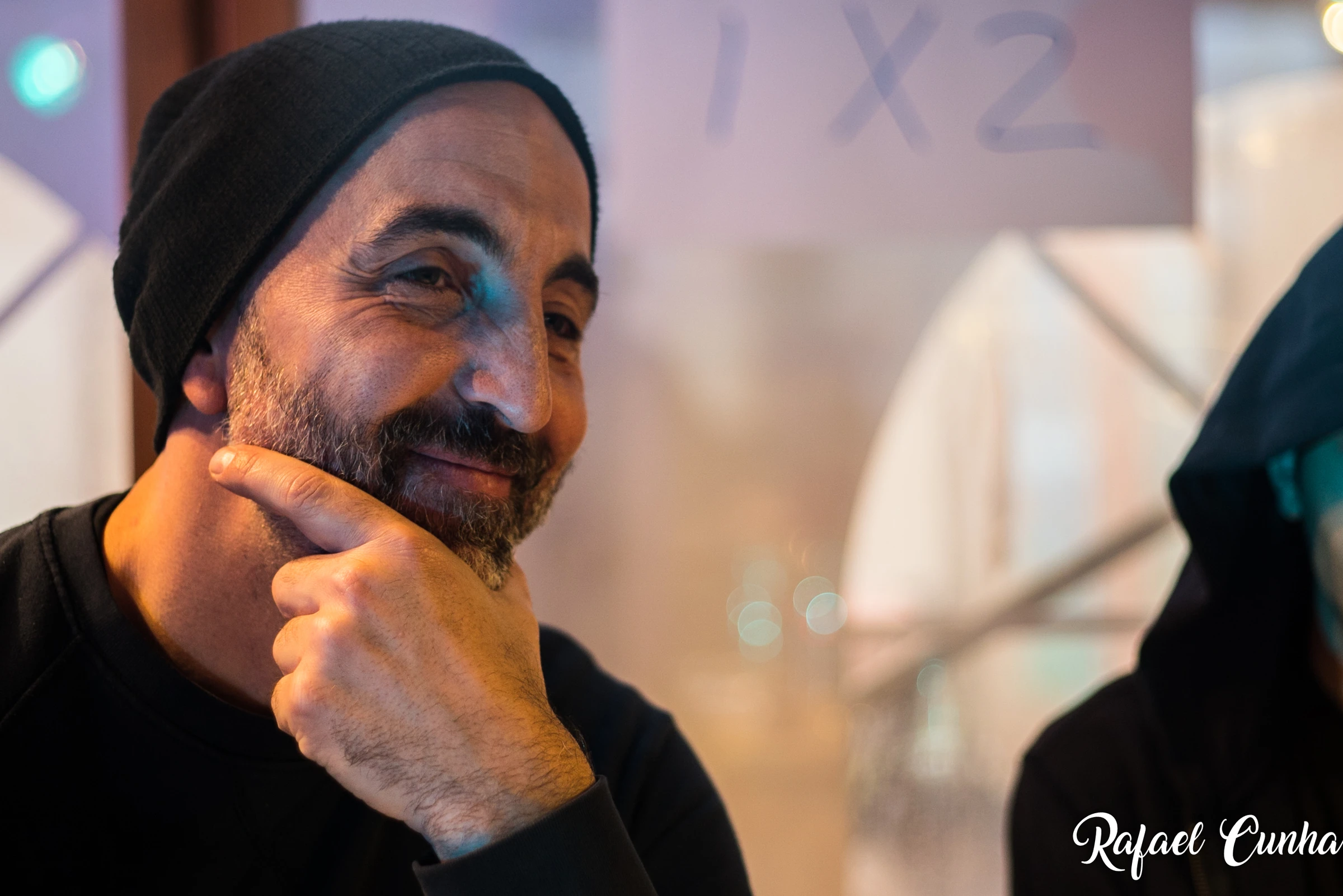 A man in a black beanie rests his hand on his chin and smiles.